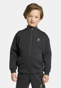 Giacca nera con zip realizzata in tessuto morbido. Presenta un colletto alto, un logo Adidas verde e polsini a costine. Abbinata a pantaloni neri coordinati.