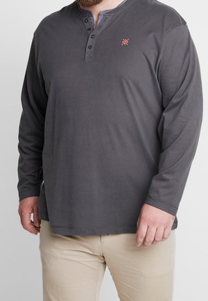 Hombre vistiendo una camisa henley de manga larga gris oscuro con botones y pantalones beige sobre un fondo liso.