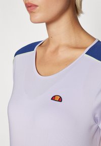 Top deportivo lila con escote redondo, acentos en los hombros azules y un logo bordado que presenta colores naranja y rojo. Textura suave.