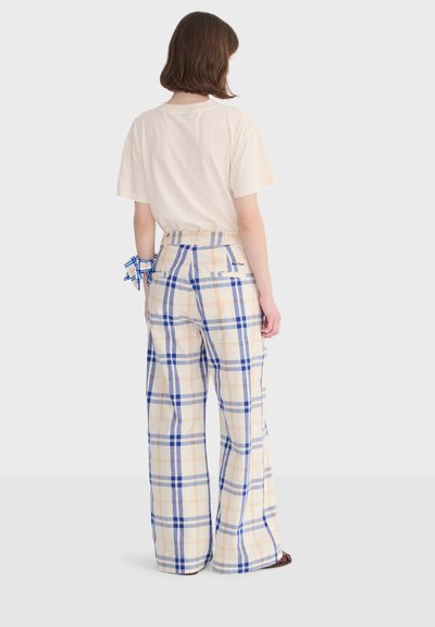 Femme debout, vue de dos, portant un t-shirt beige pâle et un pantalon large à carreaux dans des tons bleus, gris et orange, avec un foulard assorti au poignet.