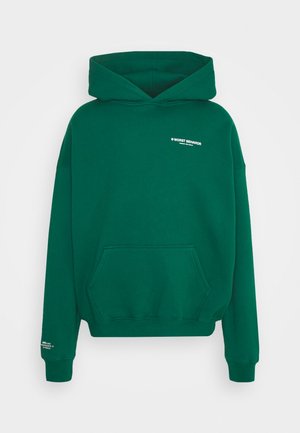 Sweat à capuche vert avec poche kangourou et petit texte blanc "WORST BEHAVIOR" sur la poitrine et la manche.