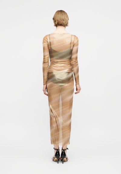 Άτομο που στέκεται με την πλάτη προς τον θεατή, φοράει μακρύ, διάφανο, μπεζ φόρεμα με σχέδια πάνω από εσώρουχα που ταιριάζουν και μαύρα σανδάλια με ψηλό τακούνι.