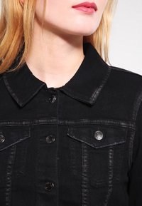 Giacca di jeans nera con colletto a punta, due tasche sul petto e chiusure con bottoni metallici. Presenta una texture sbiadita e dettagli di cucitura.