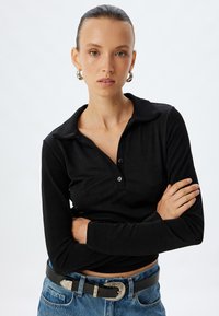 Schwarzes langärmliges Poloshirt mit Kragen und Knopfleiste mit drei Knöpfen. Kombiniert mit hochgeschnittenen blauen Jeans und einem silberverzierten Gürtel.