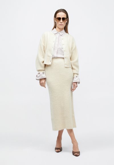 Kreemika villase ja kerge soonikkoes kardiganiga, mille all valge ja beežtriibuline särk, kombineeritud sobiva keskmise pikkusega seeliku ja pruuni kontsaga jalanõudega.
