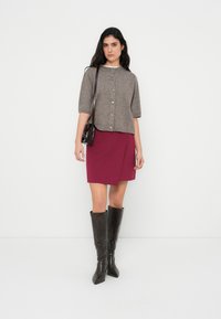 Καφέ κοντομάνικο πλεκτό ζακέτα με κουμπιά, φορεμένο πάνω από μια μπορντό φούστα τύπου φάκελου, συνδυασμένο με μαύρες δερμάτινες μπότες μέχρι το γόνατο. Μαύρη τσάντα χειρός.