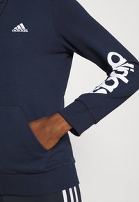 Marinblå Adidas-hoodie med vitt logotyp på ärmen, ribbade manschetter och en framficka. Tillverkad av mjukt tyg med en avslappnad passform och design.