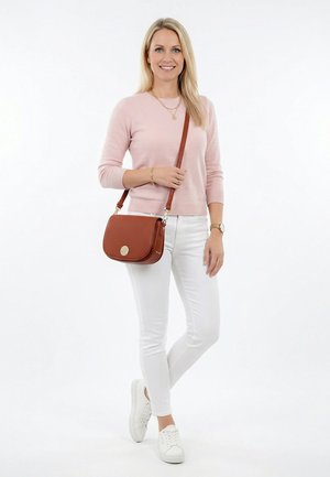 Femme blonde en pull rose, pantalon blanc et baskets blanches, souriant avec un sac bandoulière marron sur fond blanc.