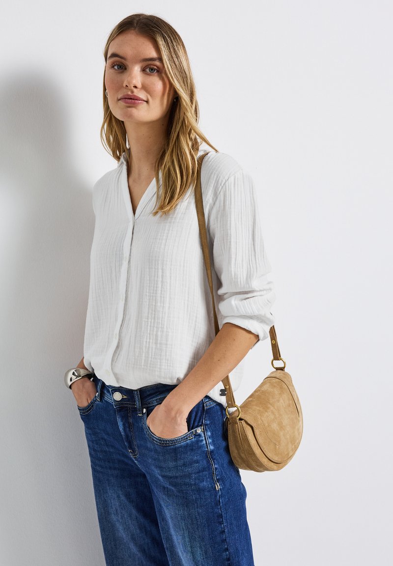 Ung kvinna med blont hår iförd vit skjorta med knappar, blå jeans, silverarmband och en beige axelväska mot en vit vägg.
