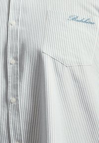 Chemise blanche et grise à rayures verticales sur le devant avec des boutons et une poche poitrine brodée du texte bleu "Badkiria".