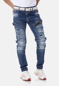 Blaue, ausgewaschene Jeans mit mehreren Rissen, versehen mit einem schwarzen Logo-Patch, kombiniert mit einem hellgrauen Nieten-Gürtel und weißen Sportschuhen.
