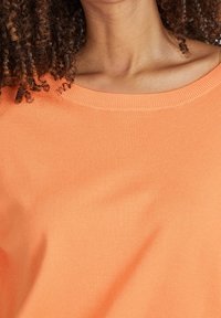 Orange lätt stickad topp med bred halsringning och ribbade kanter, med en slät textur och lätt stretch.
