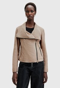 Giacca in suede beige chiaro con colletto drappeggiato, zip frontale asimmetrica e due tasche con zip. Indossata sopra un top nero.