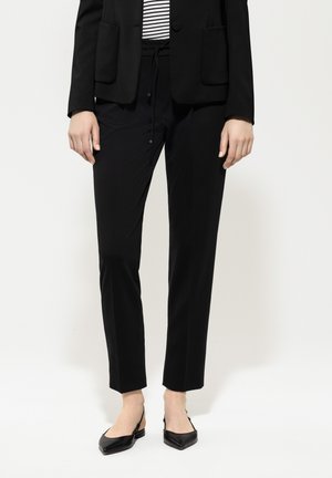 Schwarzer maßgeschneiderter Blazer mit zwei Taschen, kombiniert mit schwarzen Jogginghosen mit Kordelzug. Schwarze Slingback-Schuhe mit quadratischer Zehenform runden das Outfit ab.