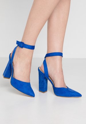 Pieds portant des chaussures à talons hauts en daim bleu vif, à bouts pointus et brides à la cheville, posés sur une surface blanche lisse.