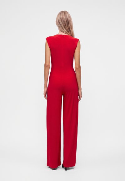 Femme aux longs cheveux blonds, de dos, portant une combinaison rouge sans manches à jambes larges et des talons noirs sur un fond uni.