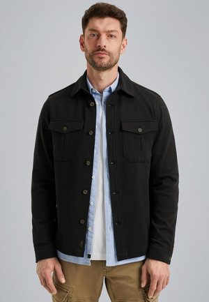 Veste noire avec deux poches poitrine, fermetures à bouton et col. Portée sur une chemise boutonnée bleu clair, associée à un pantalon kaki.