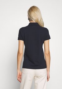 En kvinna med långt blont hår står med ryggen mot kameran, iklädd en marinblå polo shirt och ljusa byxor, framför en enkel vit bakgrund.