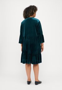 Smaragda zaļa samta kleita ar pakāpenisku volānu apakšmalu, trīs ceturtdaļu piedurknēm un gludu tekstūru. A-līnijas siluets.