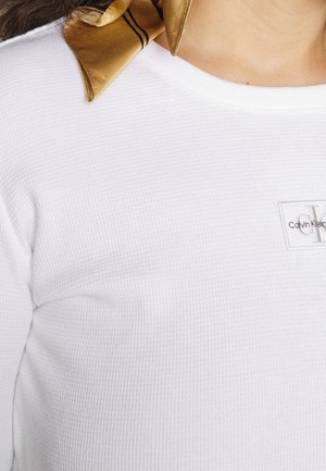 Zbliżenie na osobę noszącą fakturowaną białą koszulę Calvin Klein i złoty jedwabny szalik zawiązany wokół szyi.