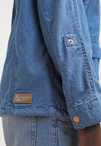 Blå denimjacka med knapplåst ärm, bröstficka och texturerat tyg. Märkbar brun etikett på fållen med logodetaljer.