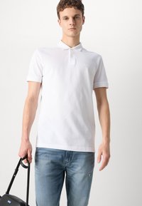 Camisa polo blanca de manga corta, con un collar y un sutil logo "BOSS" en el pecho; combinada con unos vaqueros desgastados de color azul claro y un bolso negro.