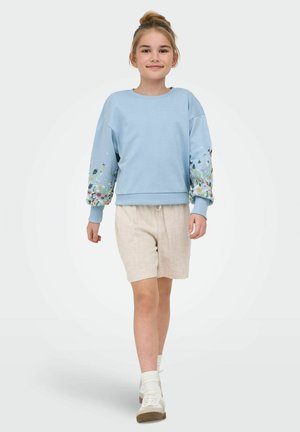 Ung pige, der går, iført en lyseblå sweatshirt med blomstrede ærmer, beige shorts, hvide sokker og hvide sneakers på en ensfarvet baggrund.