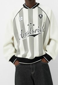 Sudadera blanca con rayas verticales negras, estampado "Amatoriale", puños y cuello de canalé negro, y dos parches de logo en el pecho.