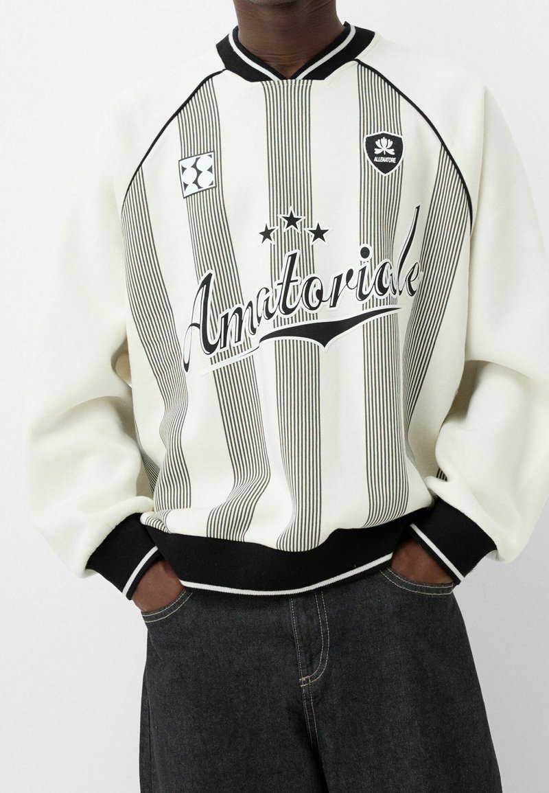 Sudadera blanca con rayas verticales negras, estampado "Amatoriale", puños y cuello de canalé negro, y dos parches de logo en el pecho.