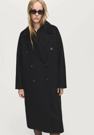 Cappotto doppiopetto in lana nera con ampi rever e bottoni, lungo fino alla caviglia, caratterizzato da una vestibilità rilassata e tasche laterali. Abbinato a occhiali da sole neri.