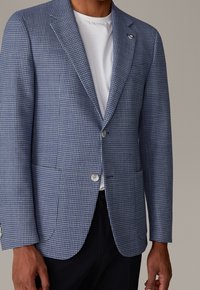 Jasnoniebieska marynarka w pepitkę z klapami z wycięciem, dwoma przednimi kieszeniami i srebrnymi guzikami, zestawiona z białym t-shirtem z okrągłym dekoltem pod spodem.