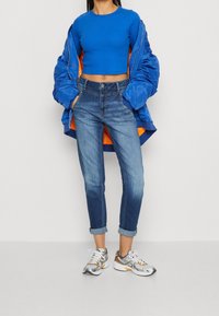 Person wearing blue cropped long-sleeve top, blue bomber jacket with orange lining, cuffed blue jeans, and silver-white sneakers.