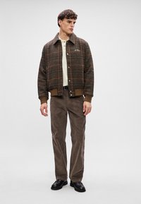 Jachetă de culoare maro, cu carouri, cu închidere prin butoane și manșete ribbed, purtată cu un tricou polo bej și pantaloni din catifea maro, completată cu pantofi negri.