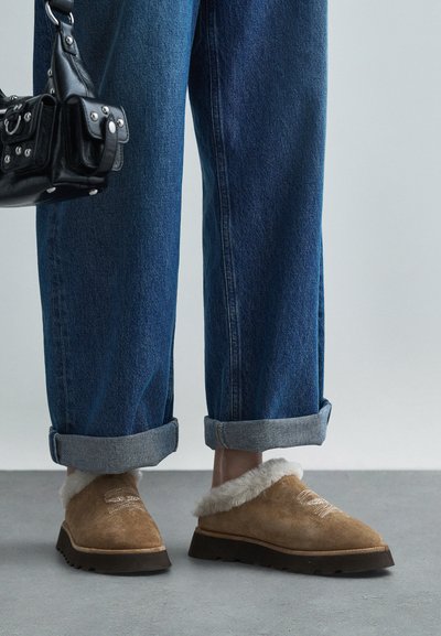 Καφέ σουέτ slip-on παπούτσια με επένδυση από γούνα, συνδυασμένα με τζιν παντελόνι σε μπλε απόχρωση με γυρισμένα μπατζάκια και μια μαύρη τσάντα, πάνω σε γκρίζα επιφάνεια.