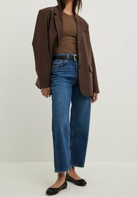 Mujer vestida con chaqueta marrón, blusa ajustada verde oliva, vaqueros de pierna ancha de altura alta en azul con cinturón negro y zapatos de ballet negros sobre fondo liso.