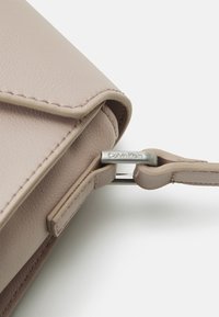 Narożnik jasnobeżowej skórzanej torebki złożony w designerski sposób, z subtelnym przeszyciem, metalowymi dodatkami i widocznym emblematem marki.