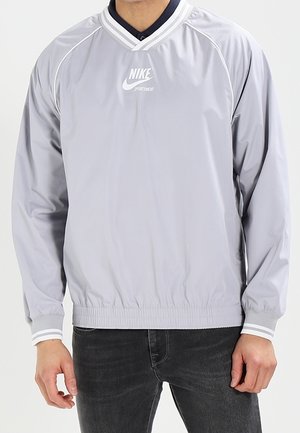 Lysegrå Nike sportspullover med hvide striber på V-hals og ærmekanter, båret med mørke jeans.