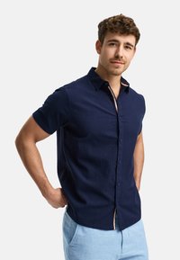 Camisa de manga corta color navy con una tela texturizada, botonera al frente y cuello interior en contraste. Llevada con pantalones azul claro, con las manos en los bolsillos.