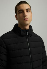 Czarna puchowa kurtka z pionowym pikowaniem, wysokim kołnierzem i zamkiem z przodu. Lekki materiał o gładkiej teksturze.