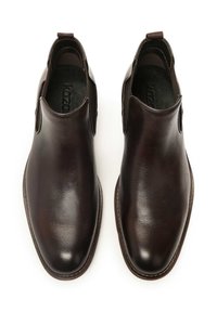 Zapatos marrones de cuero sin cordones con una textura suave, punta redondeada y dos recortes laterales. Presentan una lengüeta para facilitar el uso.
