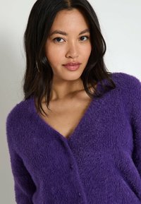 Cardigan morado y esponjoso con escote en V, que presenta una textura sutil y detalles de botones en el frente.