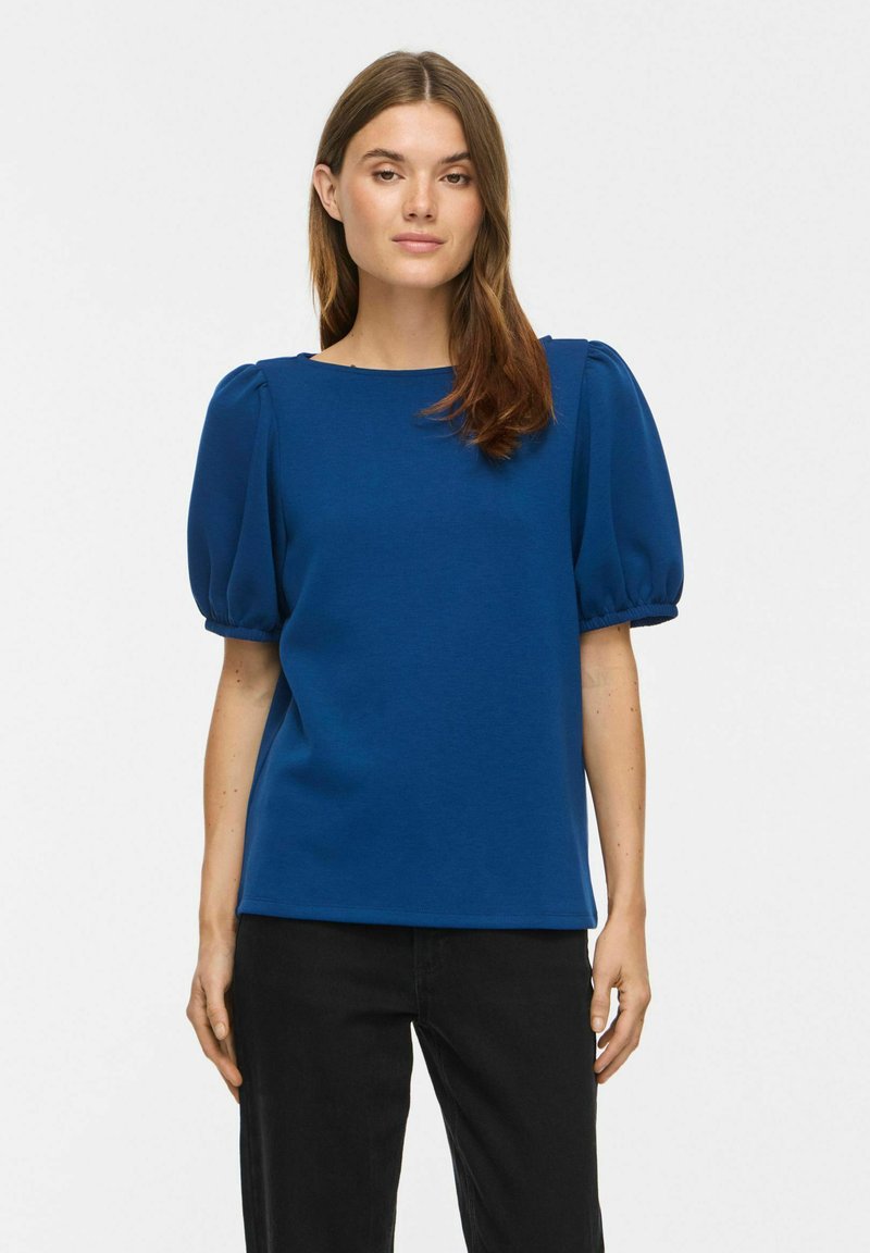 Femme aux cheveux bruns portant un haut bleu à manches courtes bouffantes et un pantalon noir, debout devant un fond blanc uni.