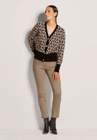 Beige gemusterter Cardigan mit V-Ausschnitt, schwarzem Besatz und fünf Knöpfen, kombiniert mit hellbeigen Hosen und schwarzen Stiefeletten.