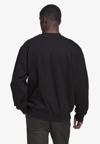 Juodas šalikas su laisvu siluetu, nusileidžiančiomis petnešomis, raištelių rankovėmis ir apvadu, matomas iš nugaros. Audinys atrodo minkštas ir tekstūrintas.