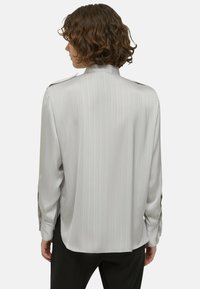 Persoon met krullend bruin haar, gekleed in een losse, lichtgrijze, verticaal gestreepte lange mouwen shirt en zwarte broek, met de rug naar de camera op een witte achtergrond.