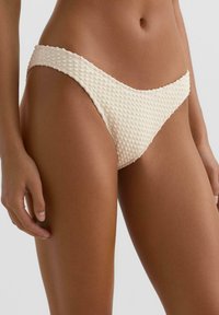 Ženska, oblečena v kremne bikini hlačke z teksturo, prikazuje trebušček in zgornji del stegen na enostavni svetli podlagi.