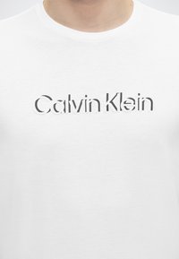 Λευκό T-shirt με στρογγυλή λαιμόκοψη και το λογότυπο "Calvin Klein" εκτυπωμένο μαύρο στο στήθος, φορεμένο από ένα άτομο με το κεφάλι κομμένο στη φωτογραφία.