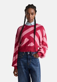 Rot und pink gemusterter Pullover mit einem cropped Design, geripptem Saum und weiten Ärmeln, kombiniert mit hoch taillierten blauen Jeans. Hellblauses Hemd darunter.
