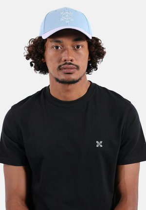 Homme aux cheveux bouclés portant une casquette bleu clair et un t-shirt noir avec un petit logo blanc brodé sur la poitrine, faisant face à l'avant.