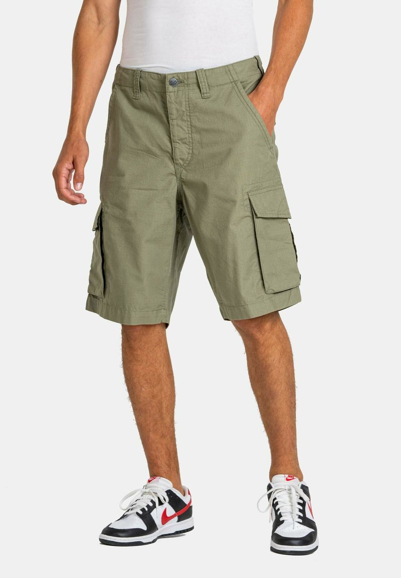 Pantalones cortos cargo verdes de tela de mezcla de algodón, con bolsillos laterales, un corte relajado y recto, combinados con zapatillas negras y blancas.